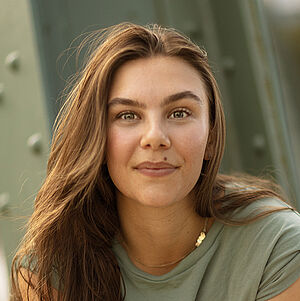 Portraitfoto von Anna Caterina Fadda. Sie sitzt mit ihrem rechten Bein auf einem stählernen grünen Gerüst. Ihre Arme sind kreuzförmig über ihr Bein gelegt. Sie hat langes braunes Haar und grinst in die Kamera. Sie trägt ein salbeifarbenes Oberteil und eine hellblaue Jeans.