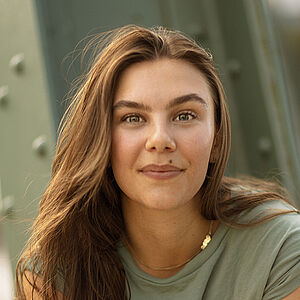 Portraitfoto von Anna Caterina Fadda. Sie sitzt mit ihrem rechten Bein auf einem stählernen grünen Gerüst. Ihre Arme sind kreuzförmig über ihr Bein gelegt. Sie hat langes braunes Haar und grinst in die Kamera. Sie trägt ein salbeifarbenes Oberteil und eine hellblaue Jeans.