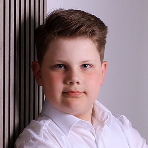 Portraitfoto von Jakob Langer. Er lehnt mit verschränkten Armen an einer Wand. Er hat blondes Haar und trägt ein weißes Hemd.