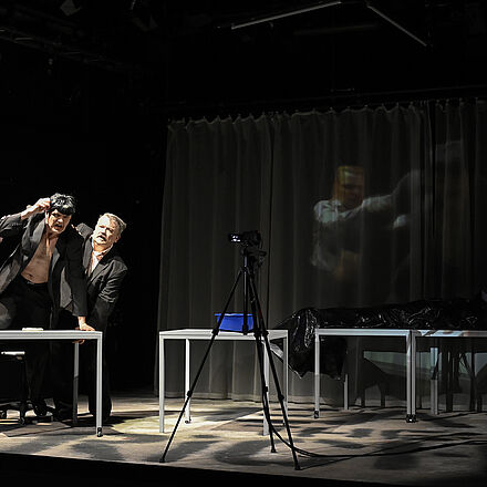 Die rechte Hälfte des Bildes liegt im Schatten. Eine leichte Projektion von drei Personen ist zu sehen. In der Mitte des Bildes steht ein Stativ. Links im Bild stehen drei Personen in einem Lichtkegel. Eine der drei Personen kniet auf einem Tisch und hält eine Hand an den Kopf.
