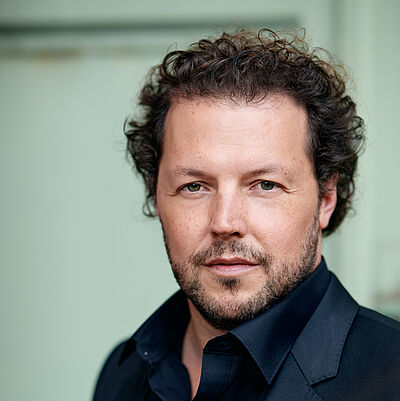 Portraitfoto von Florian Götz. Er hat kurze, braune, lockige Haare und einen Dreitagebart.  Er trägt ein schwarzes Hemd und ein schwarzes Sakko.