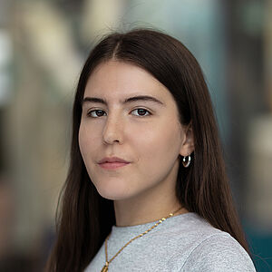 Portraitfoto von Schirin Badafaras. Sie steht seitlich da. Sie hat langes braunes Haar und trägt ein hellgraues T-Shirt sowie eine goldene Kette. Der Hintergrund ist verschwommen.