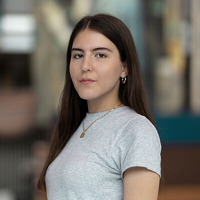 Portraitfoto von Schirin Badafaras. Sie steht seitlich da. Sie hat langes braunes Haar und trägt ein hellgraues T-Shirt sowie eine goldene Kette. Der Hintergrund ist verschwommen.