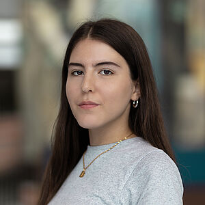Portraitfoto von Schirin Badafaras. Sie steht seitlich da. Sie hat langes braunes Haar und trägt ein hellgraues T-Shirt sowie eine goldene Kette. Der Hintergrund ist verschwommen.
