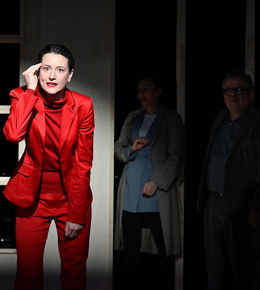 Eine Frau im roten Anzug steht im Lichtstrahl. Hinter ihr stehen vier Personen im Schatten.