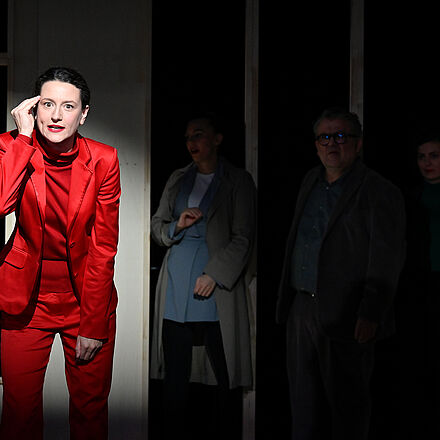 Eine Frau im roten Anzug steht im Lichtstrahl. Hinter ihr stehen vier Personen im Schatten.