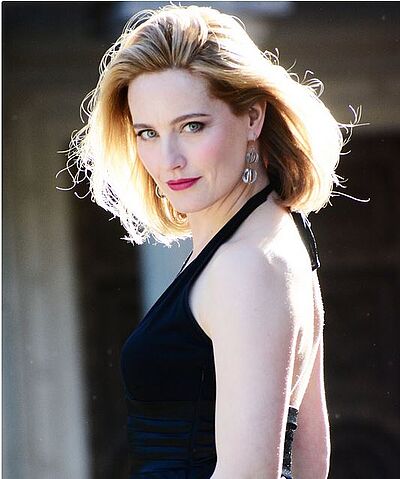 Portraitfoto von Anna Werle. Sie blickt über ihre linke Schulter. Ihr blondes schulterlanges Haar wehnt im Wind. Sie trägt ein dunkelblaues Bandeaukleid und pinken Lippenstift.