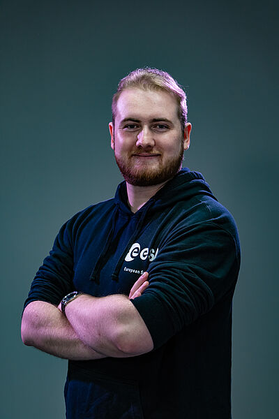 Portraitfoto von Alexander Jopke. Er steht seitlich und blickt lächelnd nach vorn. Seine Arme sind vor der Brust verschränkt. Er trägt kurzes braunes Haar, einen Vollbart und einen dunkelblauen Hoodie mit einem weißen Firmenlogo auf der linken Brust. Der Hintergrund ist grün-grau.