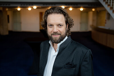 Portraitfoto von Mirko Roschkowski. Er steht in einem Theaterfoyer und blickt lächelnd in die Kamera. Er steht leicht seitlich. Sein lockiges braunes Haar fällt locker nach hinten, er trägt einen Vollbart. Er ist in einen schwarzen Anzug mit weißem Hemd gekleidet.