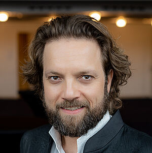 Portraitfoto von Mirko Roschkowski. Er steht in einem Theaterfoyer und blickt lächelnd in die Kamera. Er steht leicht seitlich. Sein lockiges braunes Haar fällt locker nach hinten, er trägt einen Vollbart. Er ist in einen schwarzen Anzug mit weißem Hemd gekleidet.