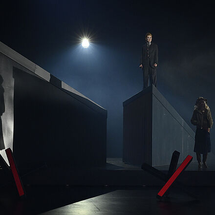 Links im Bild sieht man den Schatten einer Frau. Unten rechts im Bild ist die Frau zu sehen, die den Schatten wirft. Oben rechts steht eine andere Frau auf einer Erhöhung und blickt nach vorne. Im unteren Vordergrund stehen zwei Grenzblockaden.