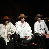 Drei Personen mit Comboyhüten sitzen am Bühnenrand. Sie tragen weiße Hemden und blicken resigniert.