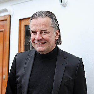 Portraitfoto von Walter Rosenberger. Er ist in einen schwarzen Rollkragenpullover und ein schwarzes Jackett gekleidet. Er lächelt. Sein graues Haar ist nach hinten gegelt.