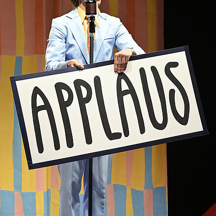 Ein Mann steht hinter einem Mikrofon und hält ein Schild mit der Aufschrift «APPLAUS» in der Hand.