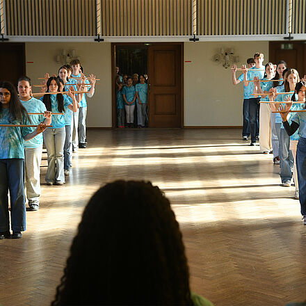 Links und rechts im Bild steht eine Reihe von Kindern. Die Kinder in den Reihen halten einen Stab in der Hand.