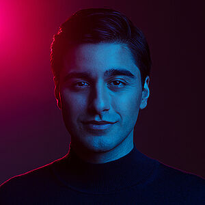 Portriatfoto von Atrin Madani. Er steht in violettem Licht und wird von hinten angeleuchtet. Er trägt einen schwarzen Pullover mit hohem Kragen. Sein Haar ist dunkel und nach hinten gestylt. Er grinst dezent.
