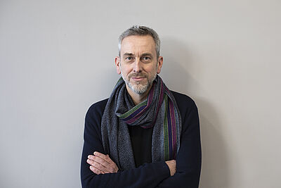 Portraitfoto von Thomas Oliver Niehaus. Er steht vor einer weißen Wand. Seine Arme sind vor der Brust verschränkt. Er trägt einen dunkelblauen Pullover, dazu einen grauen Schal mit je einem grünen, einem violetten und einem brombeerfarbenen Streifen. Sein Haar ist hellgrau und kurz, er trägt einen Bart.