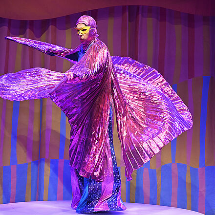 Im Mittelpunkt des Bildes steht eine Person in einem pinken/lila Gewand. Sie schwingt das Gewand in der Luft und trägt eine gelbe Maske vor den Augen.