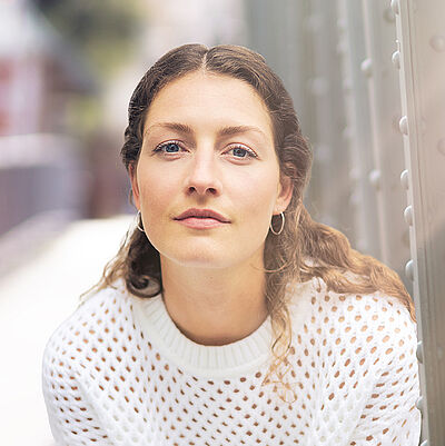 Portraitfoto von Carina Sönksen. Sie hockt in weißer Kleidung vor den Streben einer Stahlbrücke. Sie hat langes lockiges Haar.