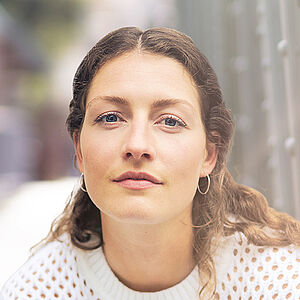 Portraitfoto von Carina Sönksen. Sie hockt in weißer Kleidung vor den Streben einer Stahlbrücke. Sie hat langes lockiges Haar. 