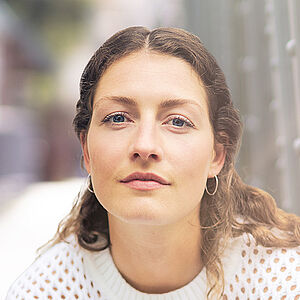Portraitfoto von Carina Sönksen. Sie hockt in weißer Kleidung vor den Streben einer Stahlbrücke. Sie hat langes lockiges Haar. 