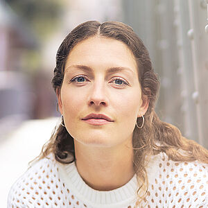 Portraitfoto von Carina Sönksen. Sie hockt in weißer Kleidung vor den Streben einer Stahlbrücke. Sie hat langes lockiges Haar. 