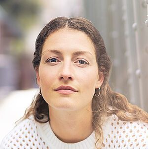 Portraitfoto von Carina Sönksen. Sie hockt in weißer Kleidung vor den Streben einer Stahlbrücke. Sie hat langes lockiges Haar. 