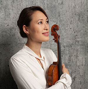 Portraitfoto von Hellen Weiß. Sie hält ihre Violine in den Armen und umgreift sie an die Brust gelehnt. Sie trägt eine weiße Bluse. Ihr Haar ist zu einem tiefen Dutt gebunden. Sie blickt nach oben. Sie steht vor einer steinernen Wand.