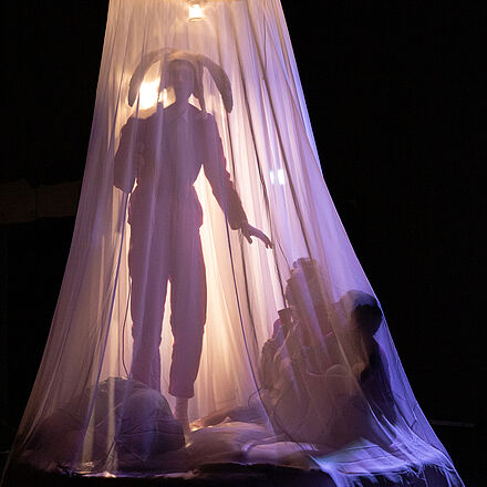Vier Personen befinden sich hinter dem Baldachin eines Himmelbetts. Sie werden von hinten mit Licht angestrahlt nur die Silhouetten der Personen sind zu sehen.
