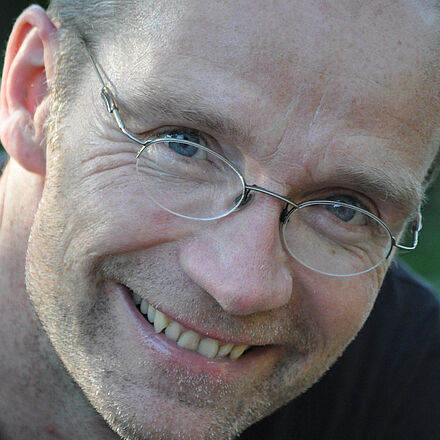 Portraitfoto von John von Düffel. Er trägt eine Brille und lächelt. Er hat kurze Haare und trägt ein dunkles Oberteil.
