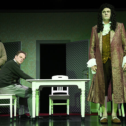 Rechts im Bild steht ein Mann in Kleidung aus der Barock-Zeit. Links im Bild sind zwei Männer. Der Mann in der Mitte sitzt an einem Tisch, der in der Mitte des Bildes steht. Im Hintergrund ist eine offenstehende Tür.