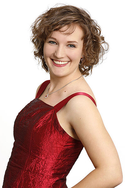 Portraitfoto von Katharina Diegritz. Sie steht seitlich und hat den linken Arm in ihre Hüfte gestützt. Ihre dunkelblunden Locken fallen ihr kinnlang ins Gesicht. Sie trägt ein rotes schimmerndes Kleid und grinst mit geöffneten roten Lippen. Der Hintergrund ist weiß.