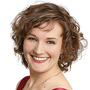 Portraitfoto von Katharina Diegritz. Sie steht seitlich und hat den linken Arm in ihre Hüfte gestützt. Ihre dunkelblonden Locken fallen ihr kinnlang ins Gesicht. Sie trägt ein rotes schimmerndes Kleid und grinst mit geöffneten roten Lippen. Der Hintergrund ist weiß.