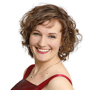 Portraitfoto von Katharina Diegritz. Sie steht seitlich und hat den linken Arm in ihre Hüfte gestützt. Ihre dunkelblonden Locken fallen ihr kinnlang ins Gesicht. Sie trägt ein rotes schimmerndes Kleid und grinst mit geöffneten roten Lippen. Der Hintergrund ist weiß.