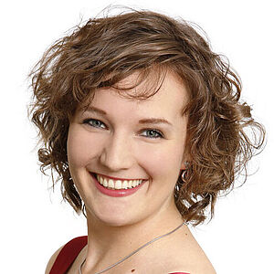Portraitfoto von Katharina Diegritz. Sie steht seitlich und hat den linken Arm in ihre Hüfte gestützt. Ihre dunkelblonden Locken fallen ihr kinnlang ins Gesicht. Sie trägt ein rotes schimmerndes Kleid und grinst mit geöffneten roten Lippen. Der Hintergrund ist weiß.