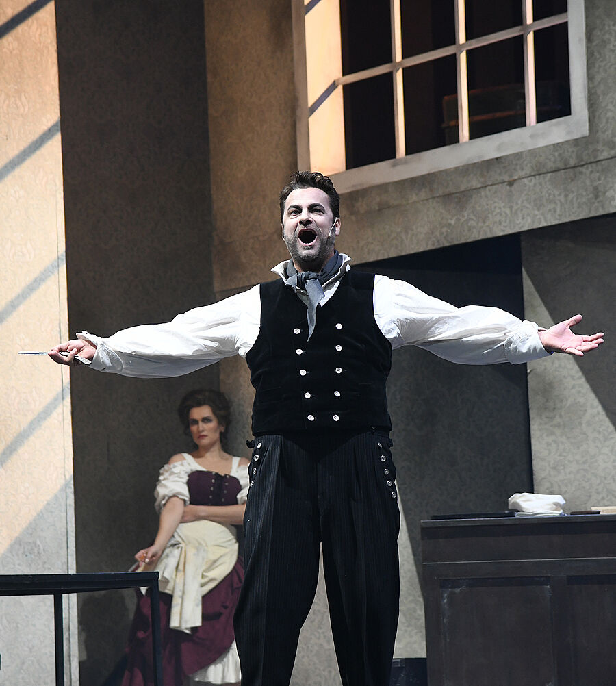 In der Mitte des Vordergrunds steht ein Mann mit offenem Mund und ausgestreckten Armen. Er hält eine Rasierklinge in der linken Hand. Im Hintergrund, hinter dem Mann, sitzt eine Frau auf einem Stuhl.