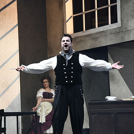 In der Mitte des Vordergrunds steht ein Mann mit offenem Mund und ausgestreckten Armen. Er hält eine Rasierklinge in der linken Hand. Im Hintergrund, hinter dem Mann, sitzt eine Frau auf einem Stuhl.