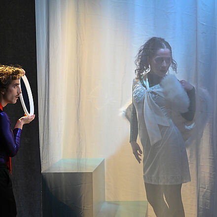 Hinter einem Vorhang steht eine Person. Sie trägt weiße Kleidung und steht auf einer Treppe. Vor dem Vorhang steht eine weitere Person. Sie trägt eine Krawatte und hält einen Spiegel in der Hand.