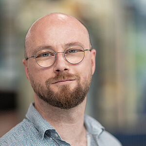 Portraitfoto von Björn Gerken. Er trägt ein hellblaues Hemd. Er hat eine Glatze, einen Vollbart und eine runde Brille auf. Der Hintergrund ist verschwommen.