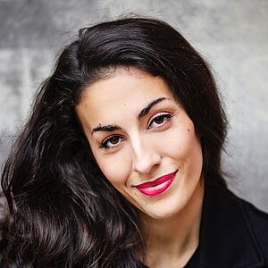 Portraitfoto von Sharon Isabelle Rupa. Sie blickt lächelnd in die Kamera. Ihr lockiges langes schwarzes Haar fällt ist über die rechte Schulter. Sie ist in einen schwarzen Blazer und ein schwarzes Oberteil gekleidet. Im Hintergrund ist eine grobe Betonmauer zu sehen.