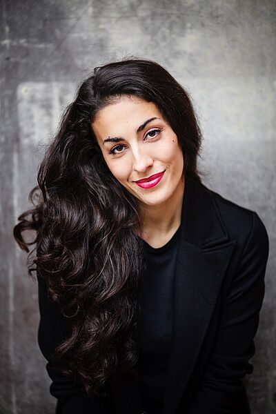 Portraitfoto von Sharon Isabelle Rupa. Sie blickt lächelnd in die Kamera. Ihr lockiges langes schwarzes Haar fällt ist über die rechte Schulter. Sie ist in einen schwarzen Blazer und ein schwarzes Oberteil gekleidet. Im Hintergrund ist eine grobe Betonmauer zu sehen.