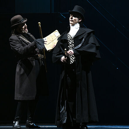 Im Mittelpunkt stehen zwei schwarz gekleidete Männer. Beide tragen Hüte und einen Gehstock. Der linke Mann hält einen Umschlag nach vorne.