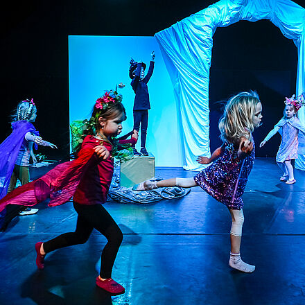 Kinder laufen und tanzen über in blumenbesetzten Kostümen auf einer blau beleuchteten Bühne.