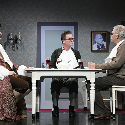 Im Mittelpunkt steht ein Tisch mit Weingläsern darauf. Am Tisch sitzen drei Männer. Der linke Mann trägt Kleidung aus der Barock-Zeit.