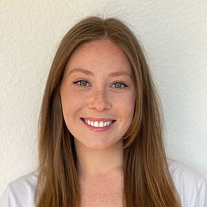 Portraitfoto von Arina Belanova. Sie lächelt mit geöffnetem Mund. Ihre dunkelblonden Haare fallen ihr auf die Brust. Sie trägt ein weißes Oberteil. Sie steht vor einem weißen Hintergrund.