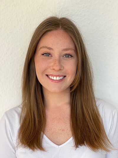 Portraitfoto von Arina Belanova. Sie lächelt mit geöffnetem Mund. Ihre dunkelblonden Haare fallen ihr auf die Brust. Sie trägt ein weißes Oberteil. Sie steht vor einem weißen Hintergrund.
