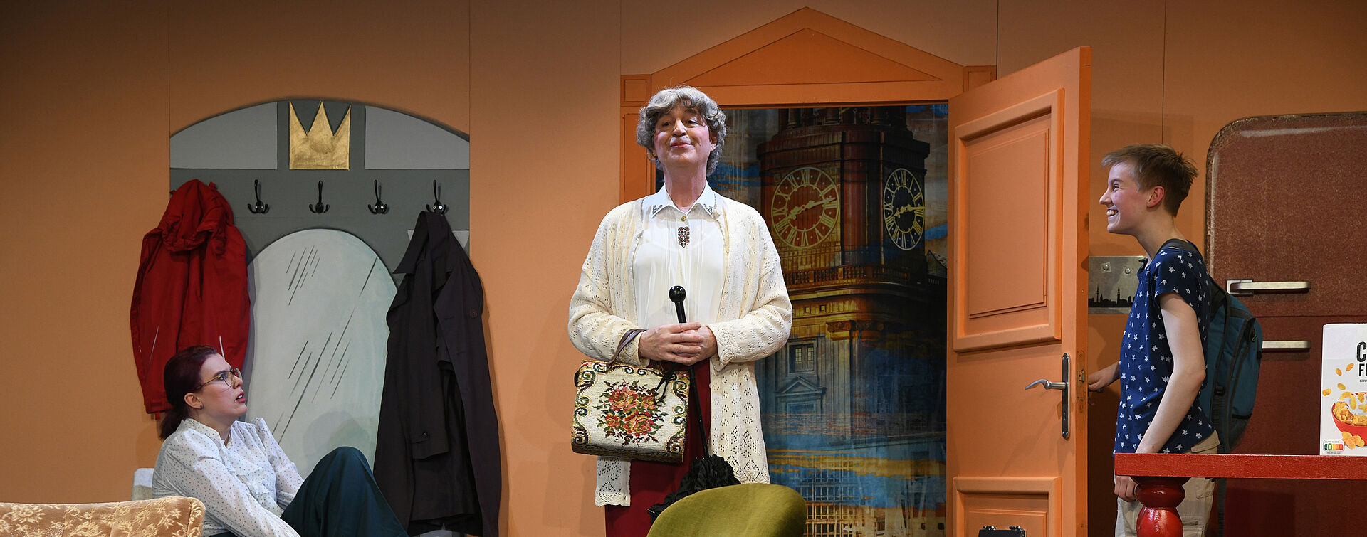 Drei Personen stehen in einem Zimmer mit orangenen Wänden. Ganz Links sitzt eine Frau mit weißer Bluse, roten Haaren und schwarzer Brille. Sie schaut überrascht auf während sie sich die Schuhe anzieht. Daneben steht ein Mann in Frauenkleidern, grauer Perücke und Schminke im Gesicht. Sie hat gerade das Zimmer betreten und lächelt. Rechts daneben steht ein Mädchen mit kurzen Haaren und einem blauen T-Shirt mit weißen Punkten. Sie trägt einen Rucksack. Sie hat gerade die Tür zum Zimmer geöffnet und den Mann hineingelassen. Sie lächelt.
