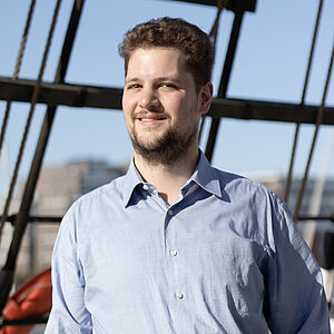Portraitfoto von Raphael Tietz. Er trägt ein hellblaues Hemd. Sein braunes Haar ist kurz und lockig. Er trägt einen gestutzten Bart. Im Hintergrund sind Holzstreben und Seile zu sehen. Dahinter ist der blaue Himmel.