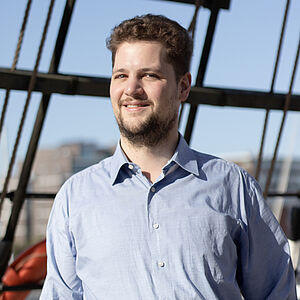Portraitfoto von Raphael Tietz. Er trägt ein hellblaues Hemd. Sein braunes Haar ist kurz und lockig. Er trägt einen gestutzten Bart. Im Hintergrund sind Holzstreben und Seile zu sehen. Da-hinter ist der blaue Himmel.