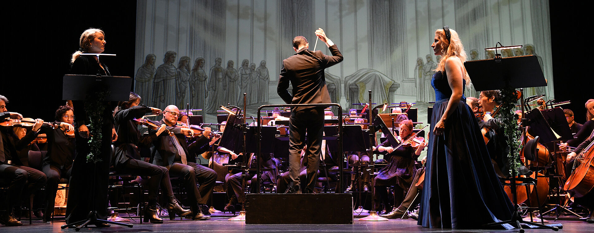 Im Vordergrund stehen zwei Personen, die sich ansehen. Dahinter ist das Orchester zu sehen. Als Hintergrundbild sind gezeichnete, graue Fenster erkennbar. 